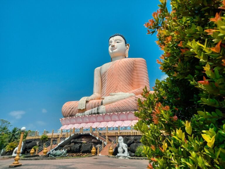 A large Buddha statue under a clear sky at a temple in Bentota, Sri Lanka, surrounded by greenery.